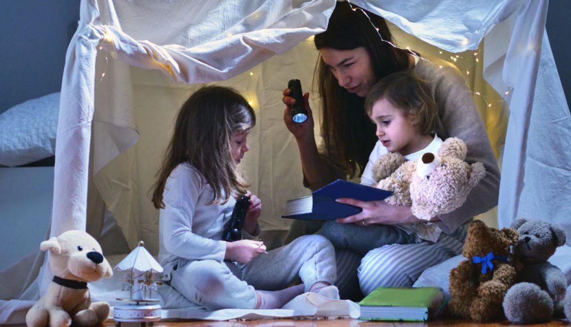 A mother sits with her daughters in a home made tent indoors.