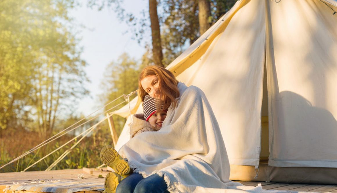 Young mother embracing her happy kid with a blanket while sitting near canvas bell tent in the forest