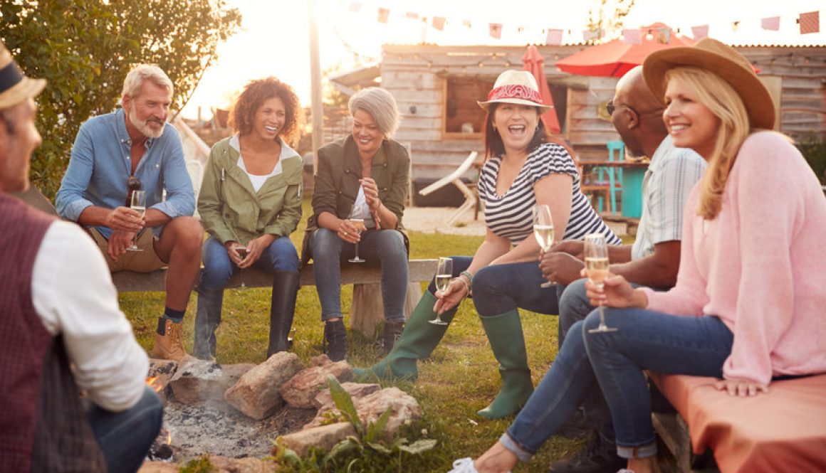 Group of mature friends drinking alcohol, sitting by fire at campsite.