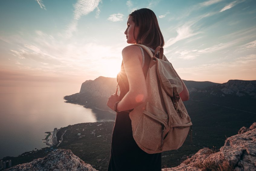 Young woman with backpack enjoys the view of the mountains and the sea at sunset.