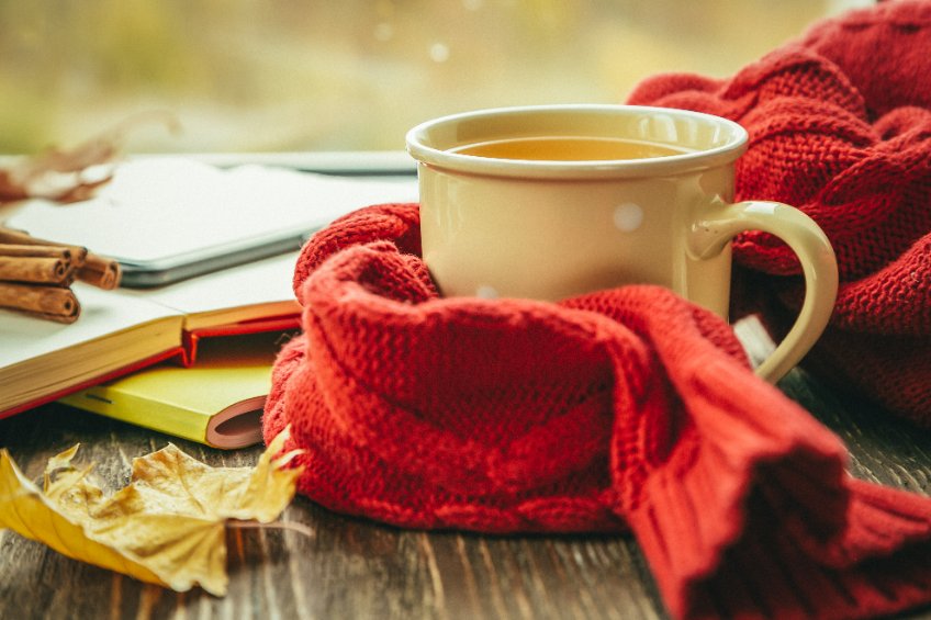 A mug of tea with a red scarf around it next to Autumnal leaves.